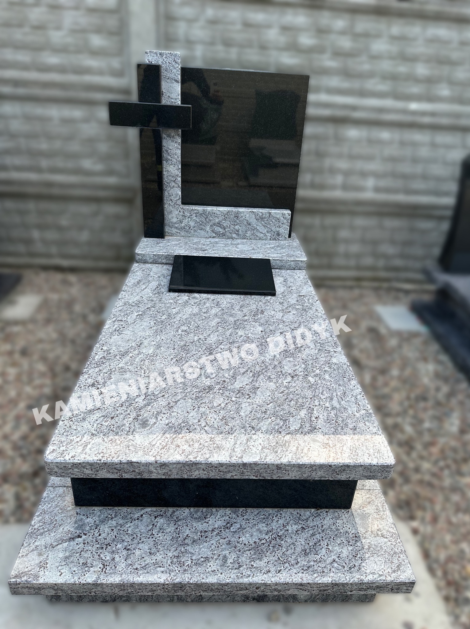 Jasnoszary, dwupoziomowy pomnik z granitu z czarnym krzyżem i tabliczką, widoczne ziarno kamienia i napis 'Kamieniarstwo Didyk'.