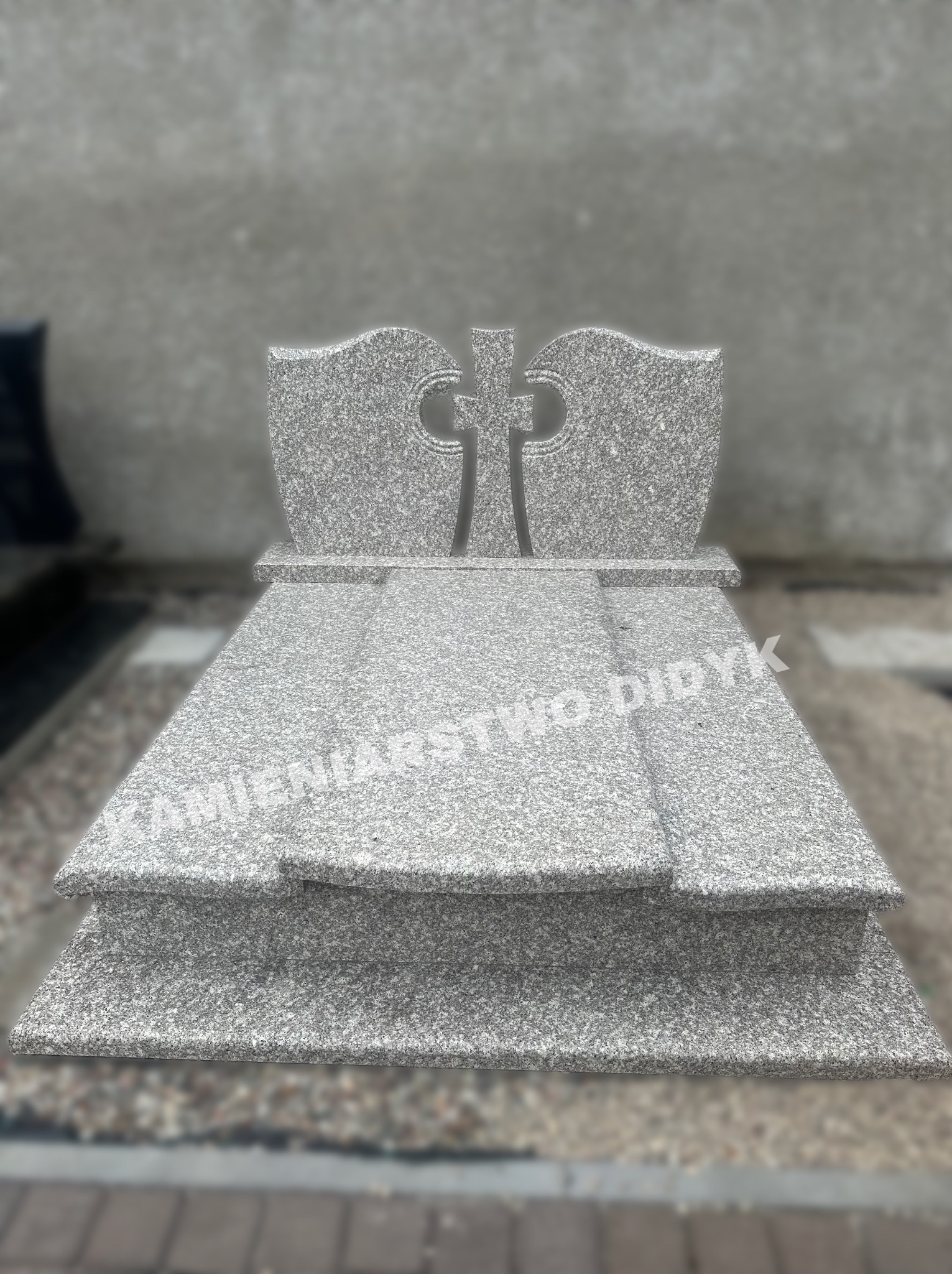 Szary, granitowy pomnik z krzyżem i napisem 'KAMIENIARSTWO DIDYK', ustawiony na kostce brukowej.