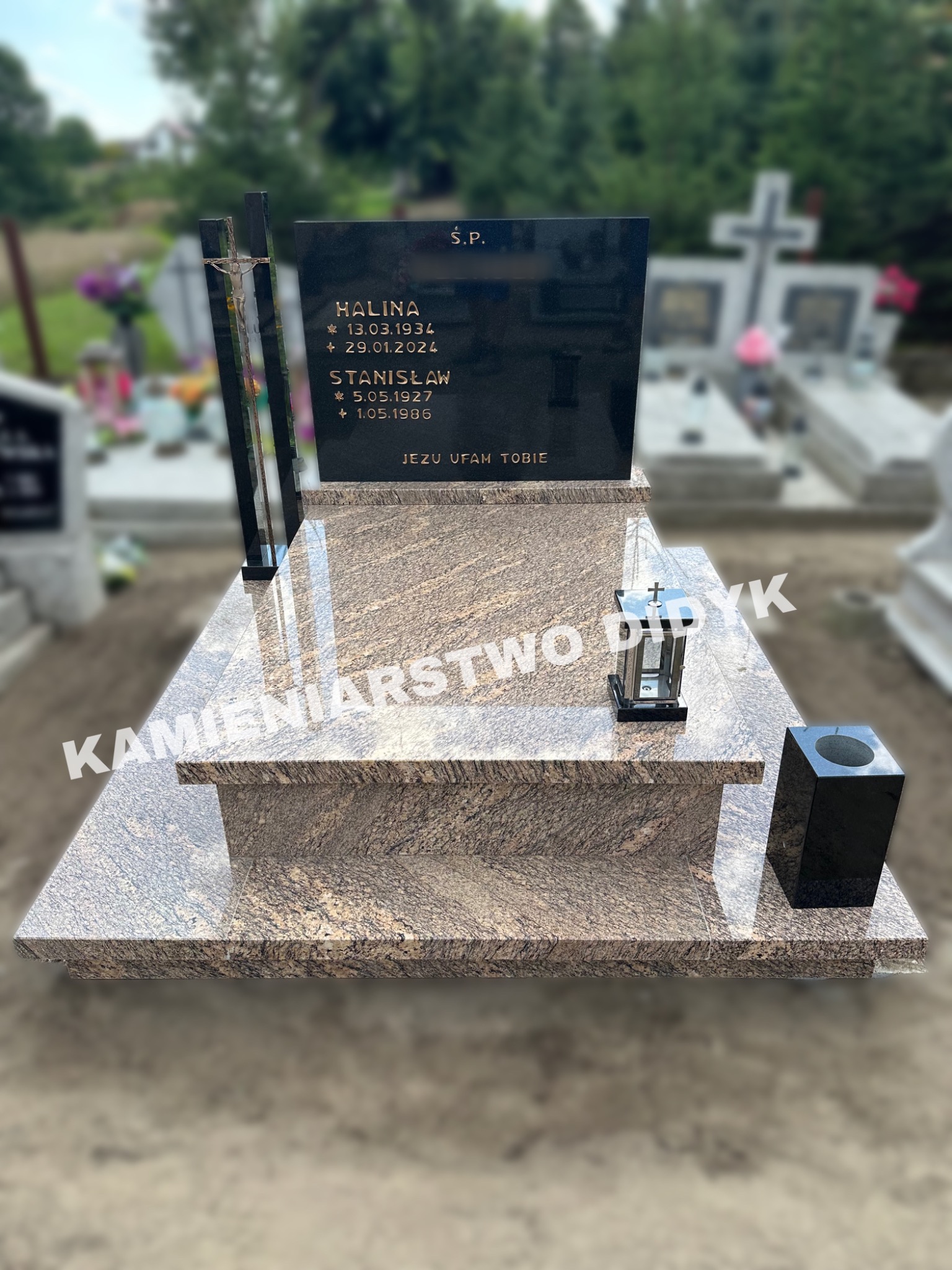 Granitowy pomnik nagrobny z czarną tablicą inskrypcyjną, metalowym krzyżem i lampionem, na tle innych nagrobków i zieleni cmentarza.