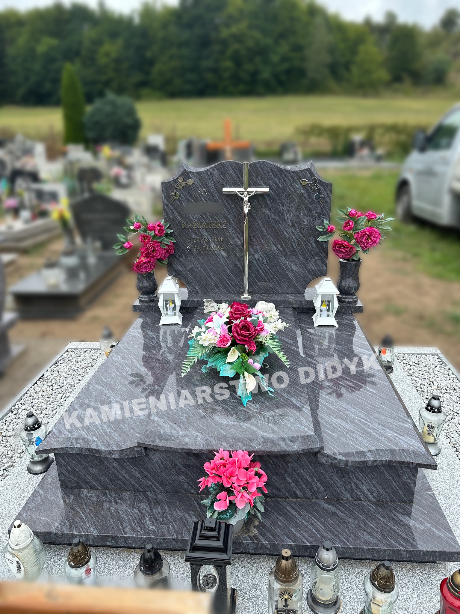 Granitowy nagrobek z krzyżem i wygrawerowanym napisem 'Kamieniarstwo Didyk', ozdobiony kwiatami i lampionami, na tle cmentarza.