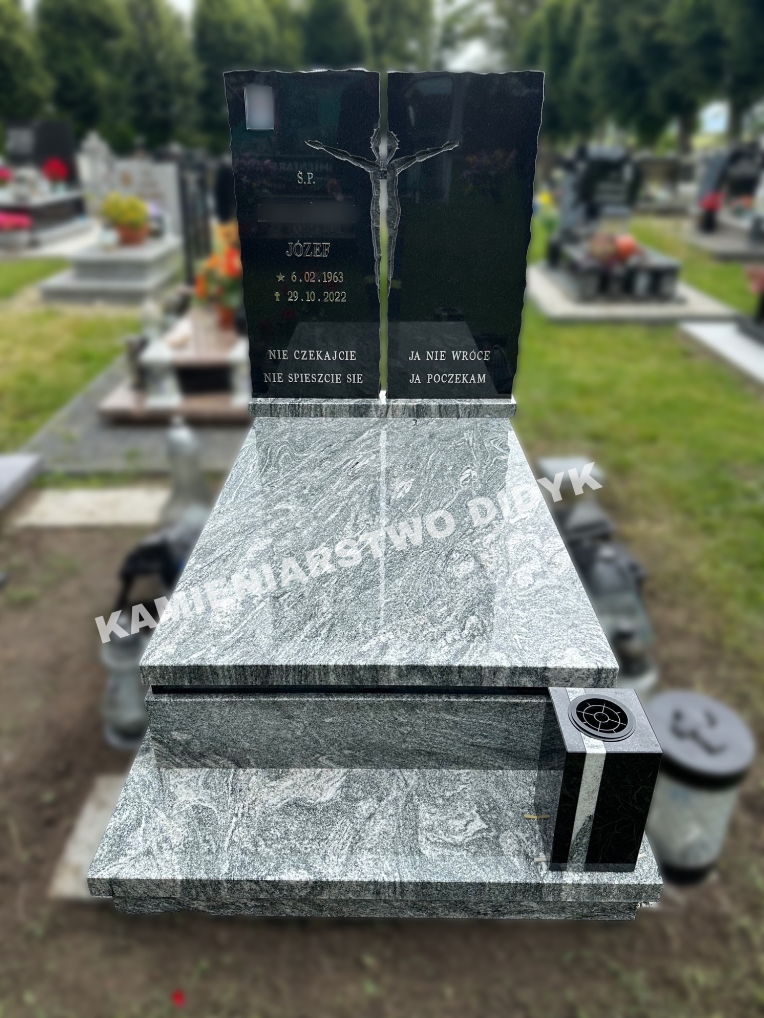 Dwuczęściowy, granitowy pomnik z wyrytym krucyfiksem i inskrypcjami na cmentarzu, z czarnym lampionem po prawej stronie i innymi nagrobkami w tle.