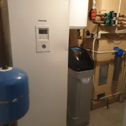 Biała pompa ciepła Panasonic Aquarea, obok zmiękczacz wody Water Technic i niebieski zbiornik wyrównawczy, instalacja hydrauliczna z rurami z tworzywa na tle beżowej ściany.