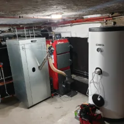 Piwnica z zainstalowanym kotłem na paliwo stałe marki Biadala, zasobnikiem Kospel, naczyniem wzbiorczym i rozbudowaną instalacją hydrauliczną pod sufitem.