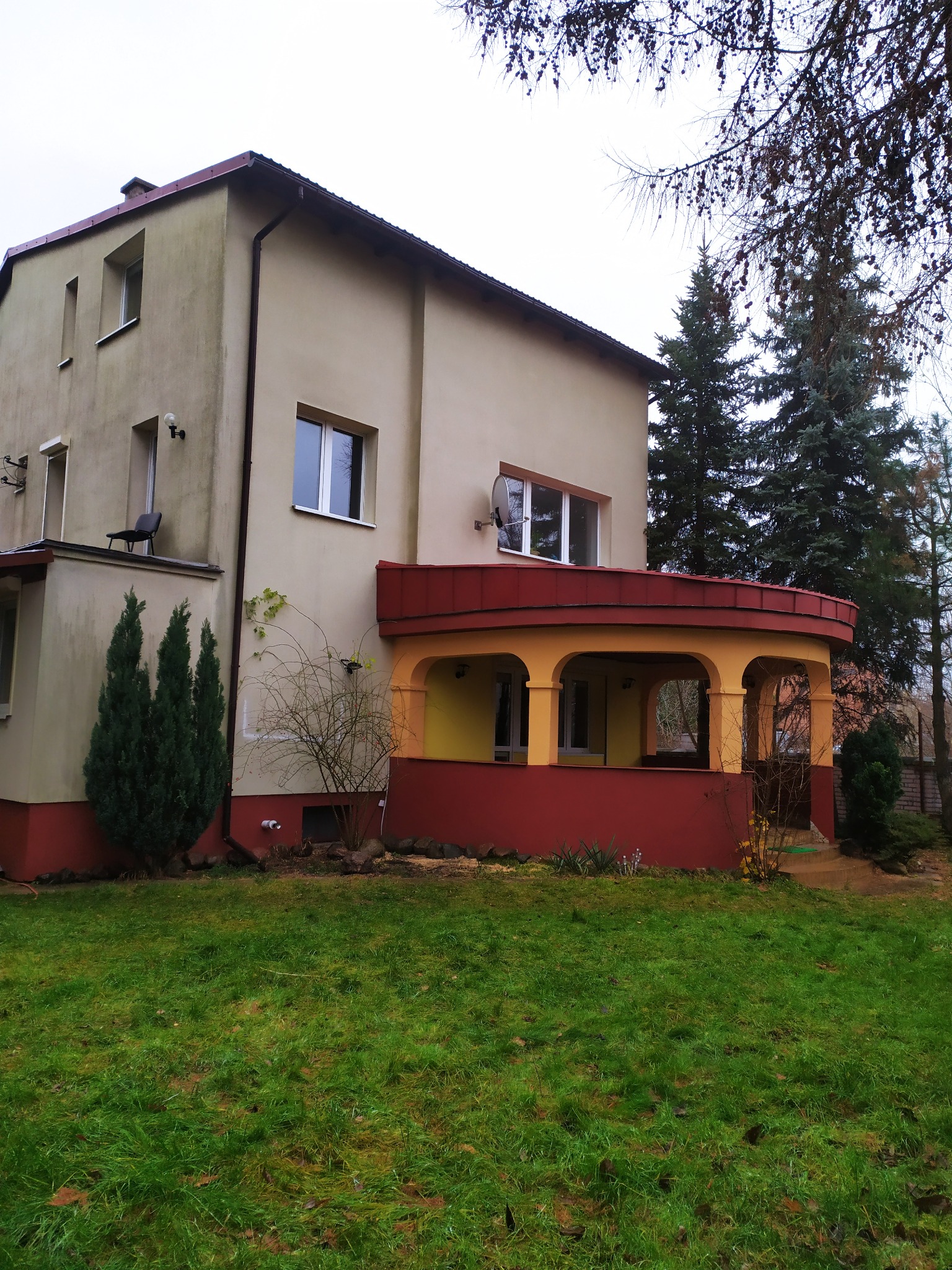 Dwupiętrowy dom z czerwonym dachem i rynnami, widok z trawnika w pochmurny dzień.
