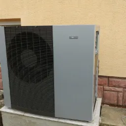 Zewnętrzna jednostka szarej pompy ciepła NIBE F2120 zainstalowana przy ścianie budynku w Dębicy, widoczny fragment okna i elewacji z cegieł.