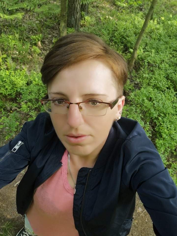 Kobieta w okularach, ubrana w różową koszulkę i granatową kurtkę, stoi w lesie, patrząc w dół na aparat.