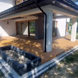 Zadaszony taras z drewna, z betonową donicą wypełnioną piaskiem, przylegający do nowoczesnego domu z dużymi oknami przesuwnymi.