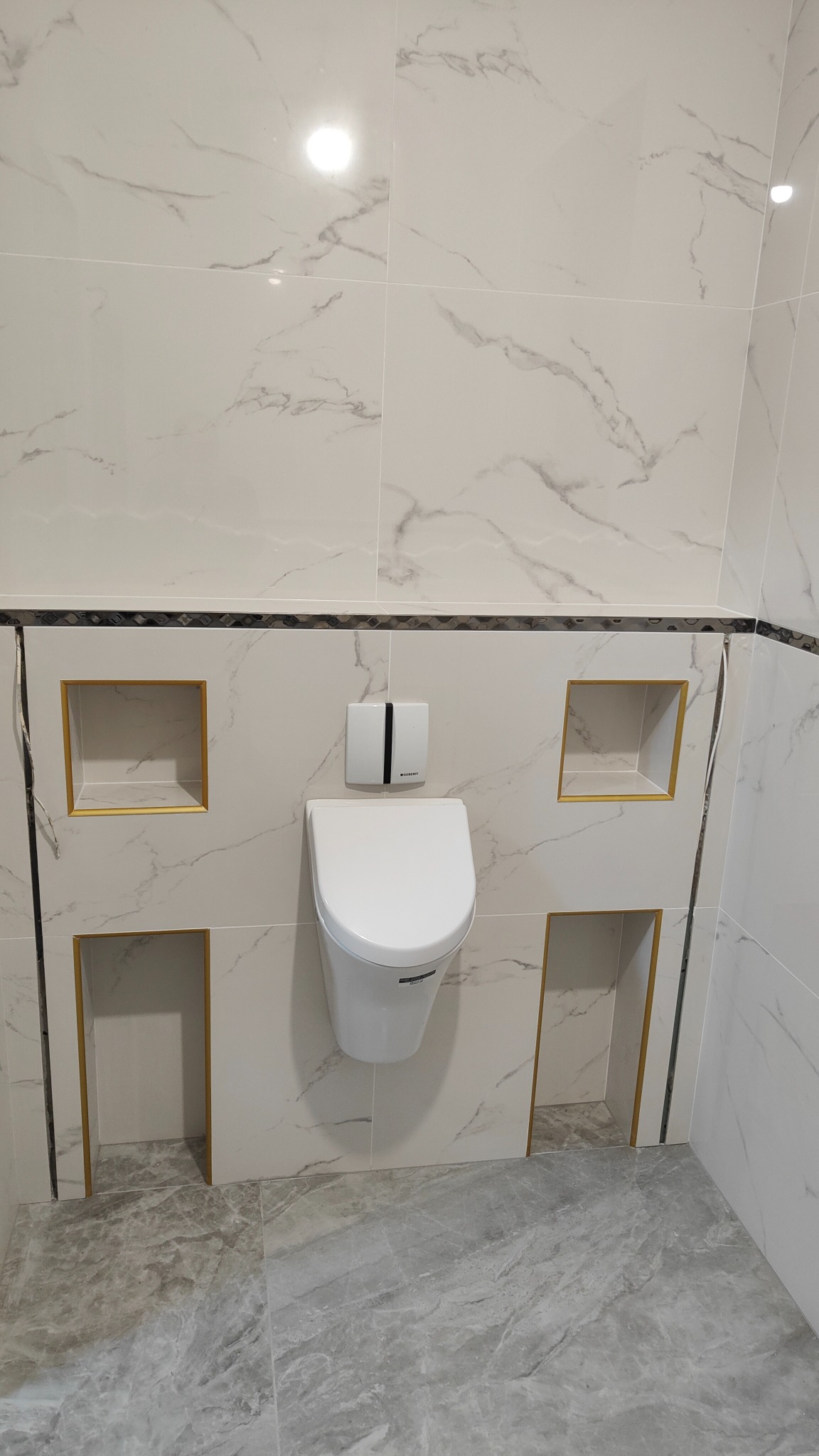 Nowoczesna toaleta podwieszana z białymi płytkami marmurkowymi i szarymi płytkami podłogowymi, wnęki z dekoracyjnym złotym obramowaniem w ścianie.