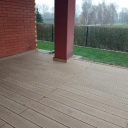 Nowo wykończony taras z desek kompozytowych w odcieniach beżu, przylegający do ceglanego budynku z czerwoną kolumną, na tle zielonego trawnika i krzewów.