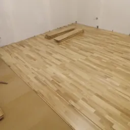 Układanie jasnej drewnianej podłogi parkietowej w pokoju, widoczne deski, podkład i ściany z wystającymi przewodami elektrycznymi.