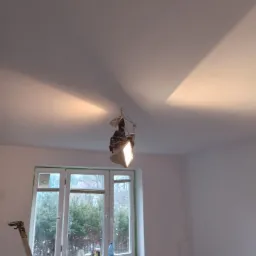 Pok&oacute;j po malowaniu, widoczne świeżo pomalowane ściany i sufit, okno zabezpieczone taśmą malarską, drabina oparta o ścianę, na parapecie butelki i akcesoria malarskie, lampa wisząca na środku sufitu.