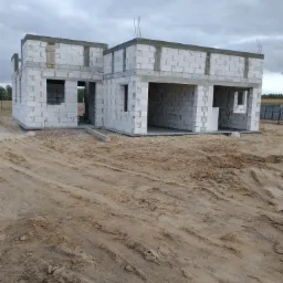 Surowy stan parterowego domu z betonu komórkowego w trakcie budowy, widoczne otwory okienne i drzwiowe, teren wokół przygotowany pod dalsze prace.