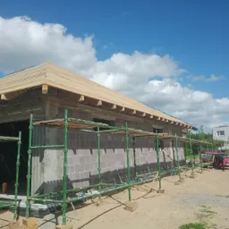 Budowa domu z pustaków: widok na konstrukcję dachu z drewnianym deskowaniem, rusztowania wzdłuż ściany, błękitne niebo z chmurami w tle.