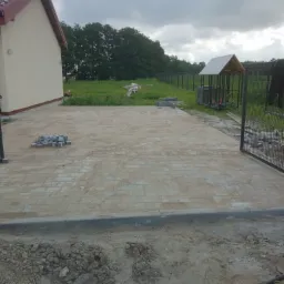 Teren przed domem z częściowo ułożoną kostką brukową, widoczne krawężniki i brama wjazdowa, w tle zieleń i ogrodzenie.