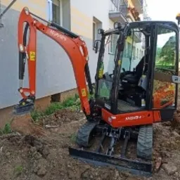 Pomarańczowa mini koparka Kubota wykonuje wykop w ziemi przy budynku mieszkalnym, widoczne ślady świeżo naruszonej ziemi i roślinność.