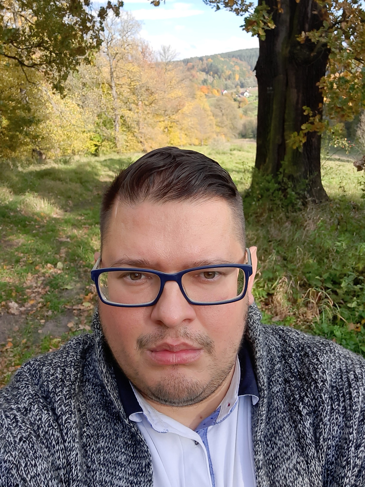 Mężczyzna w okularach z niebieskimi oprawkami, w swetrze na tle jesiennego krajobrazu z drzewami i wzgórzami w oddali.