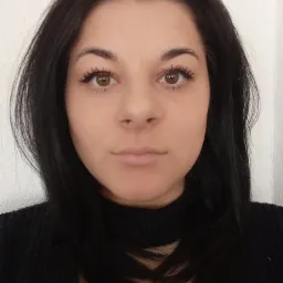 Portret kobiety z ciemnymi włosami i brązowymi oczami na tle białej ściany, ubrana w czarny sweter z golfem.