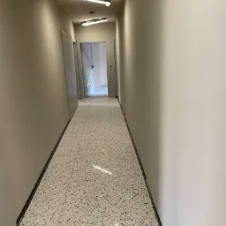 Korytarz z posadzką lastryko w odcieniach szarości i beżu, ściany w kolorze złamanej bieli, sufit z trzema lampami w ochronnych foliach, drzwi widoczne w głębi korytarza.