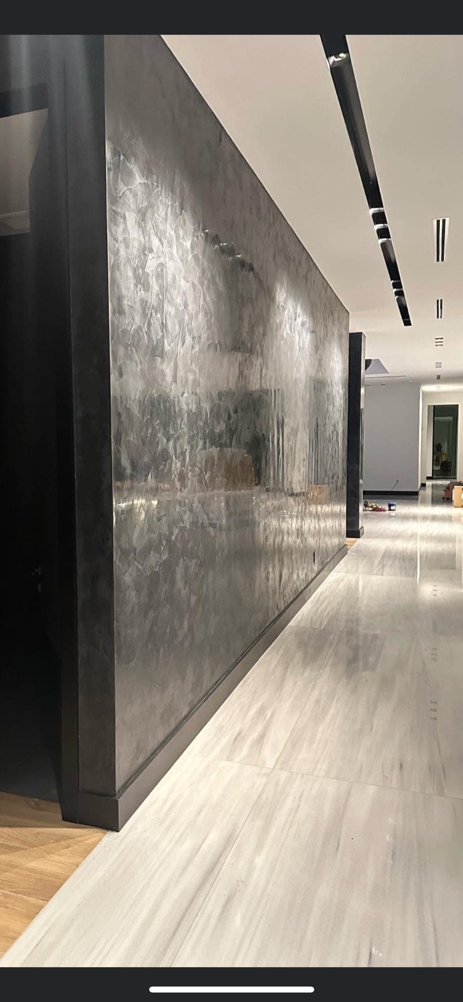 Długa ściana w korytarzu pokryta dekoracyjną, srebrzystą farbą z efektem połysku, ułożona w nieregularny wzór, z ciemną listwą przypodłogową i marmurową podłogą.