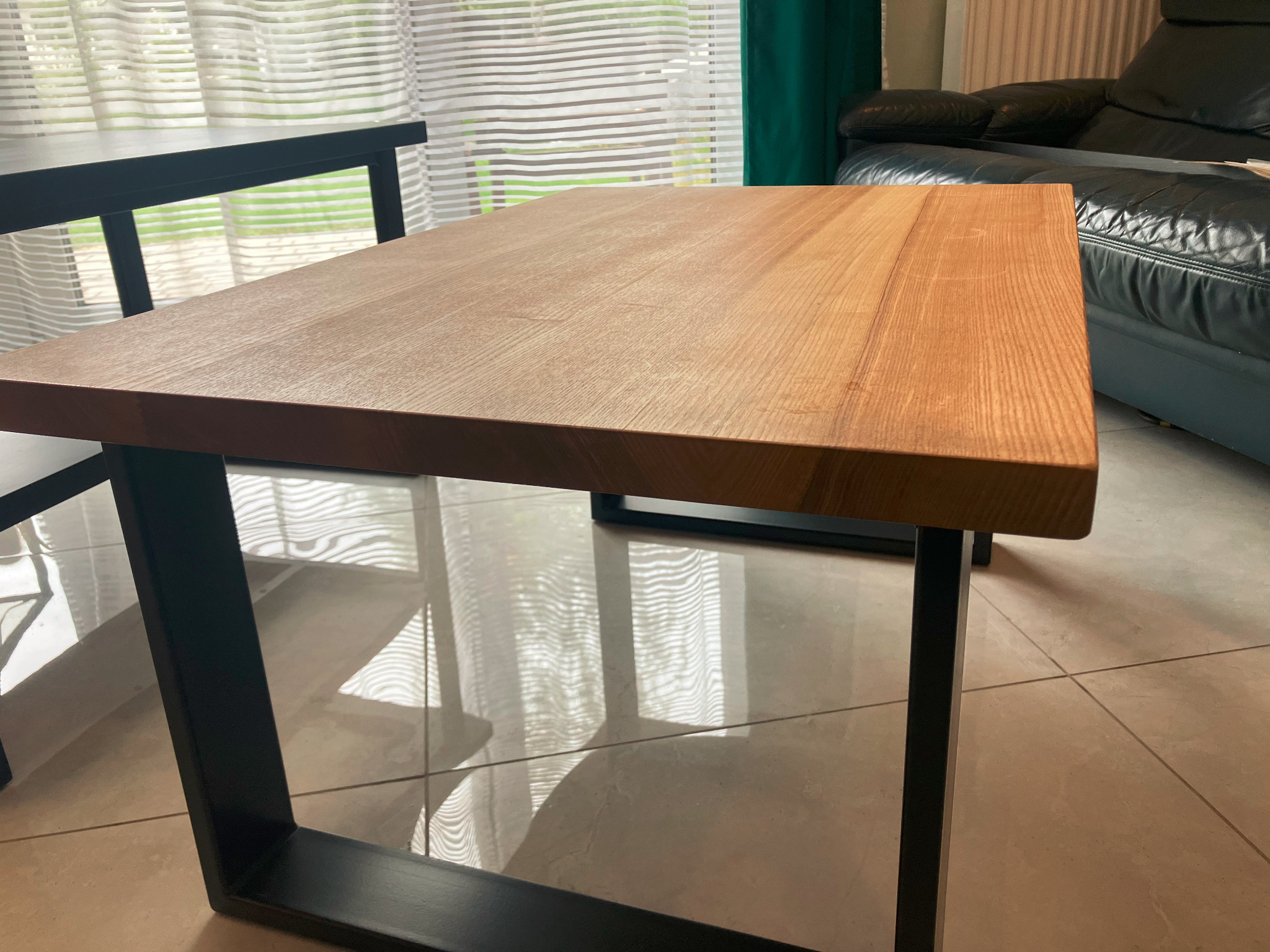 Stół kawowy z blatem z jasnego drewna i czarnymi, metalowymi nogami o kwadratowym przekroju, odbijający się w polerowanej podłodze z płytek, na tle okna z żaluzjami i skórzanej sofy.