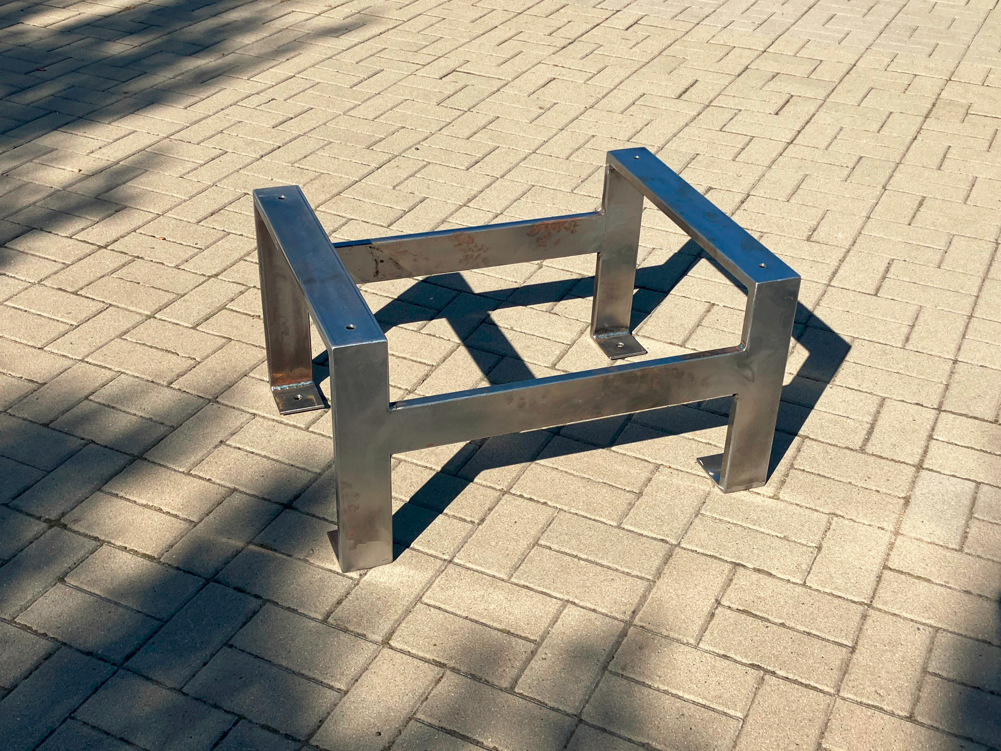 Surowa, spawana konstrukcja stalowa w kształcie podstawy stołu, widoczna na brukowanej powierzchni w słoneczny dzień, z widocznymi śladami spawania i otworami montażowymi.