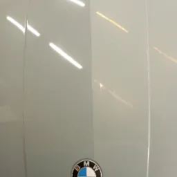 Prawa strona maski samochodu BMW po polerowaniu, widoczna różnica w połysku w porównaniu do niepolerowanej lewej strony. Logo BMW na dole maski.