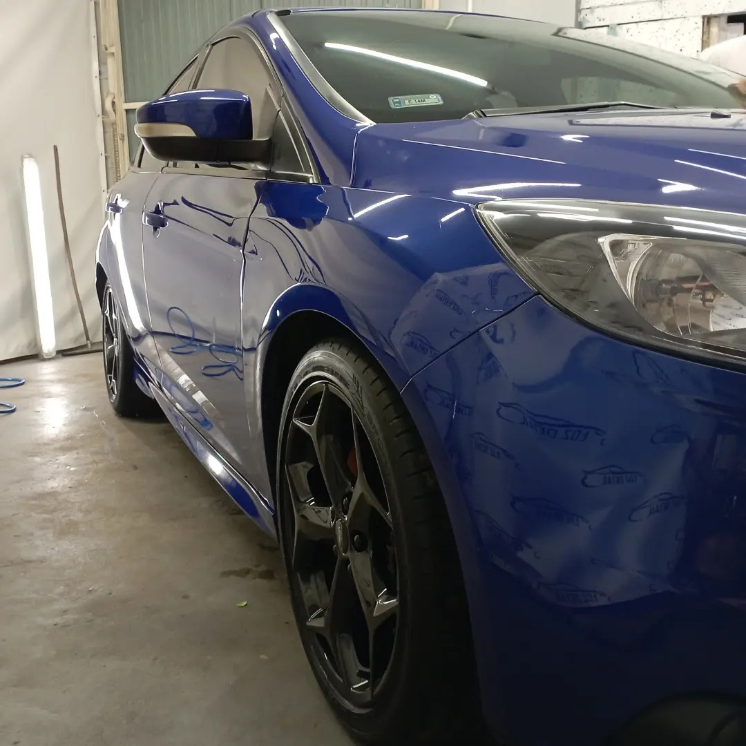 Błyszczący, niebieski samochód po umyciu, widoczny z bliska, z czarnymi felgami, na tle ściany myjni.