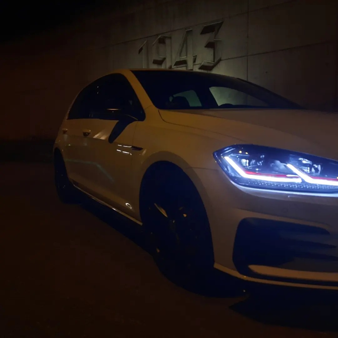 Biały samochód osobowy z włączonymi światłami, zaparkowany w tunelu z wyrytym rokiem 1943 na ścianie.