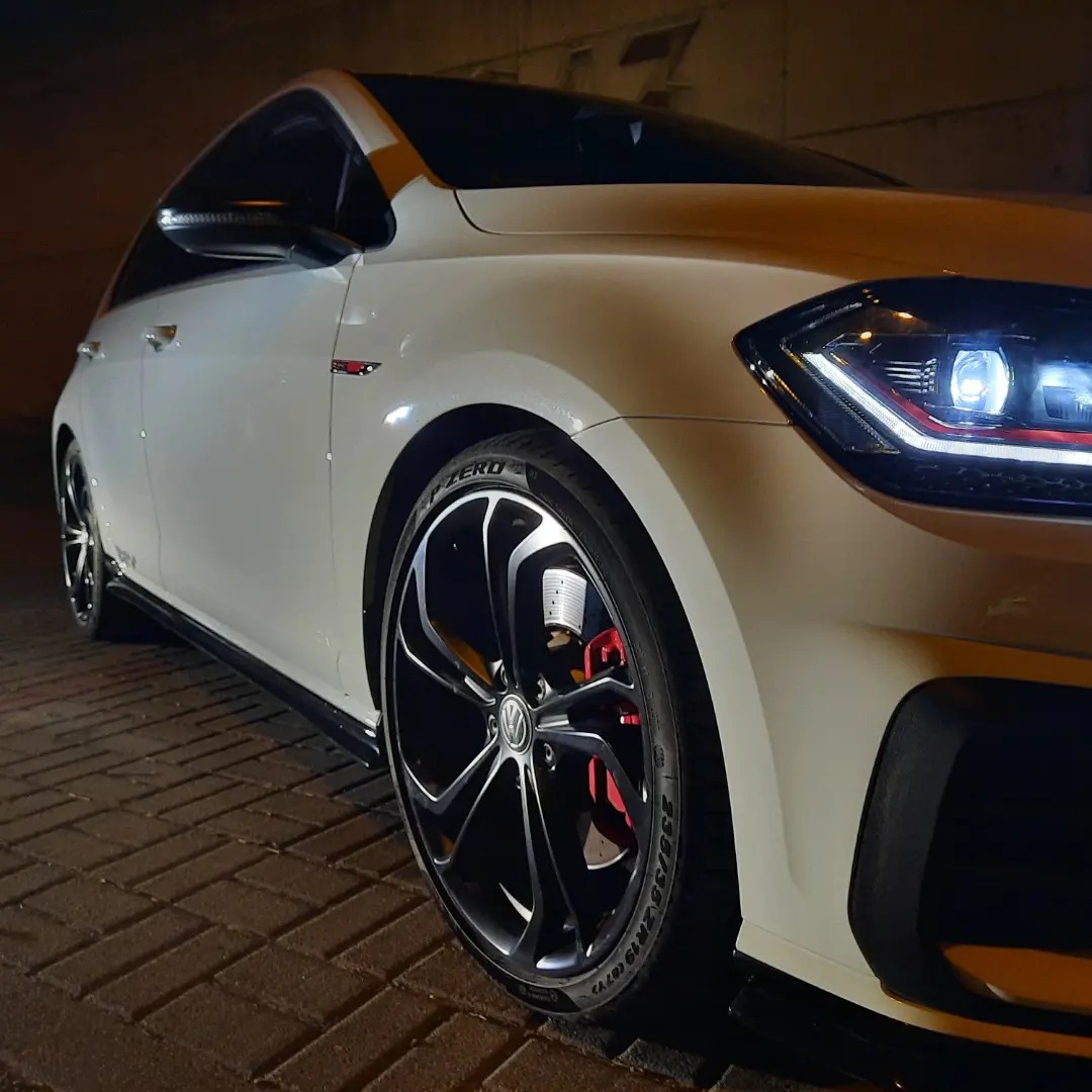 Biały samochód VW Golf GTI z czarnymi felgami i czerwonymi zaciskami hamulcowymi, sfotografowany pod niskim kątem na tle ceglanej nawierzchni.