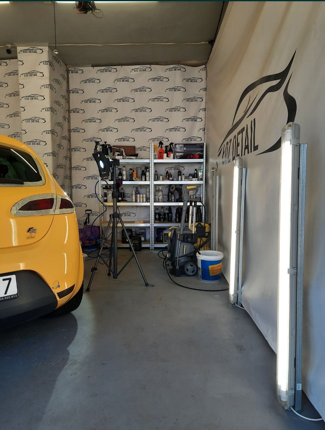 Żółty samochód w studio detailingowym z oświetleniem LED, regałem z preparatami i myjką ciśnieniową, widoczne logo firmy na ścianie