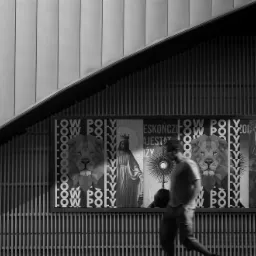 Czarno-białe zdjęcie przedstawia plakat z grafikami low poly, w tym lwa i postaci religijne, umieszczony na ścianie budynku z pionowymi listwami; przechodzący mężczyzna jest lekko rozmazany w ruchu.