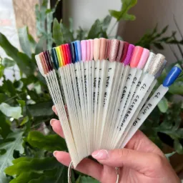 Paleta lakierów do paznokci w kształcie wachlarza, prezentująca różne kolory i wykończenia, trzymana w dłoni na tle zielonych roślin.