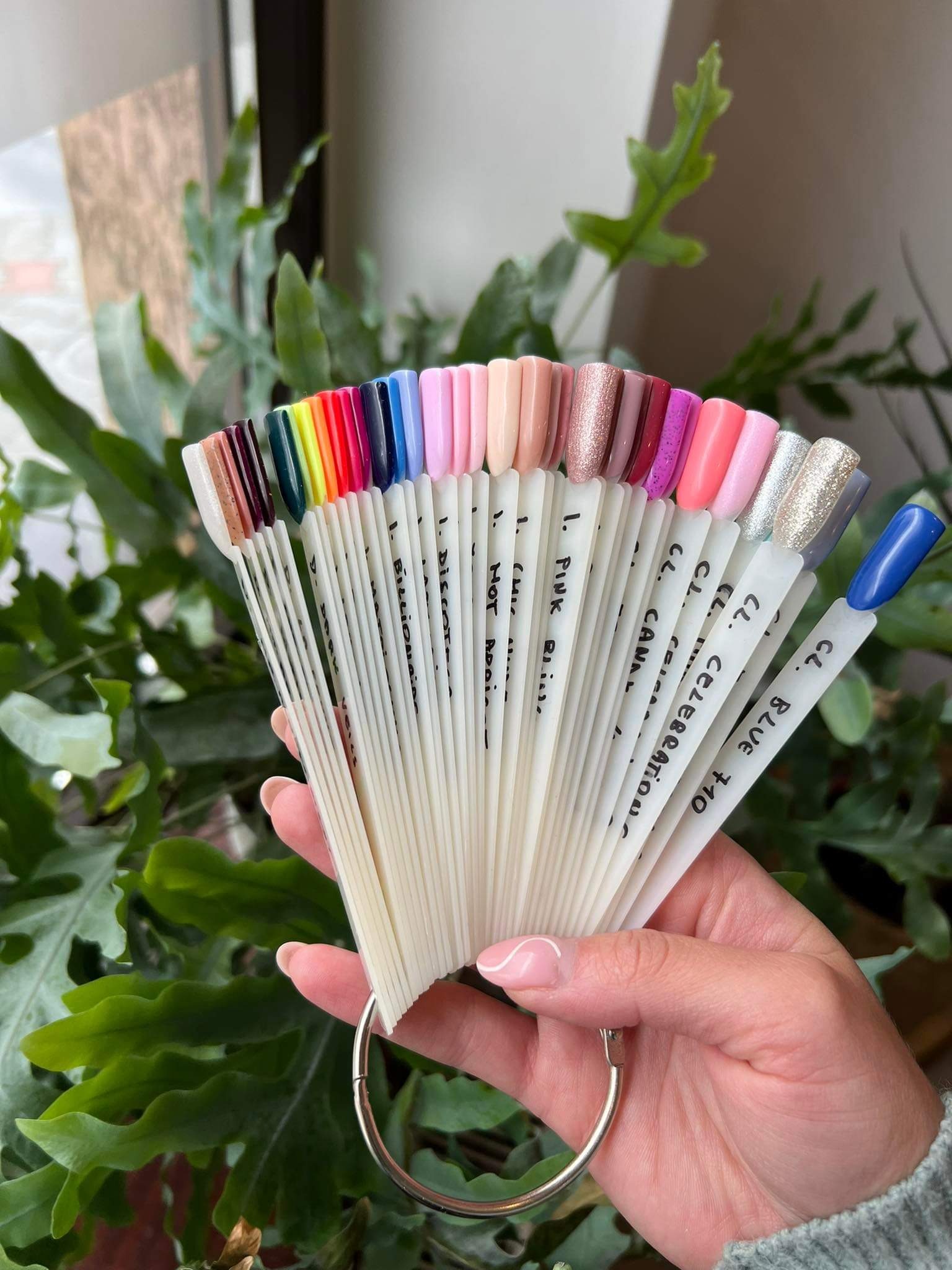 Paleta lakierów do paznokci w kształcie wachlarza, prezentująca różne kolory i wykończenia, trzymana w dłoni na tle zielonych roślin.