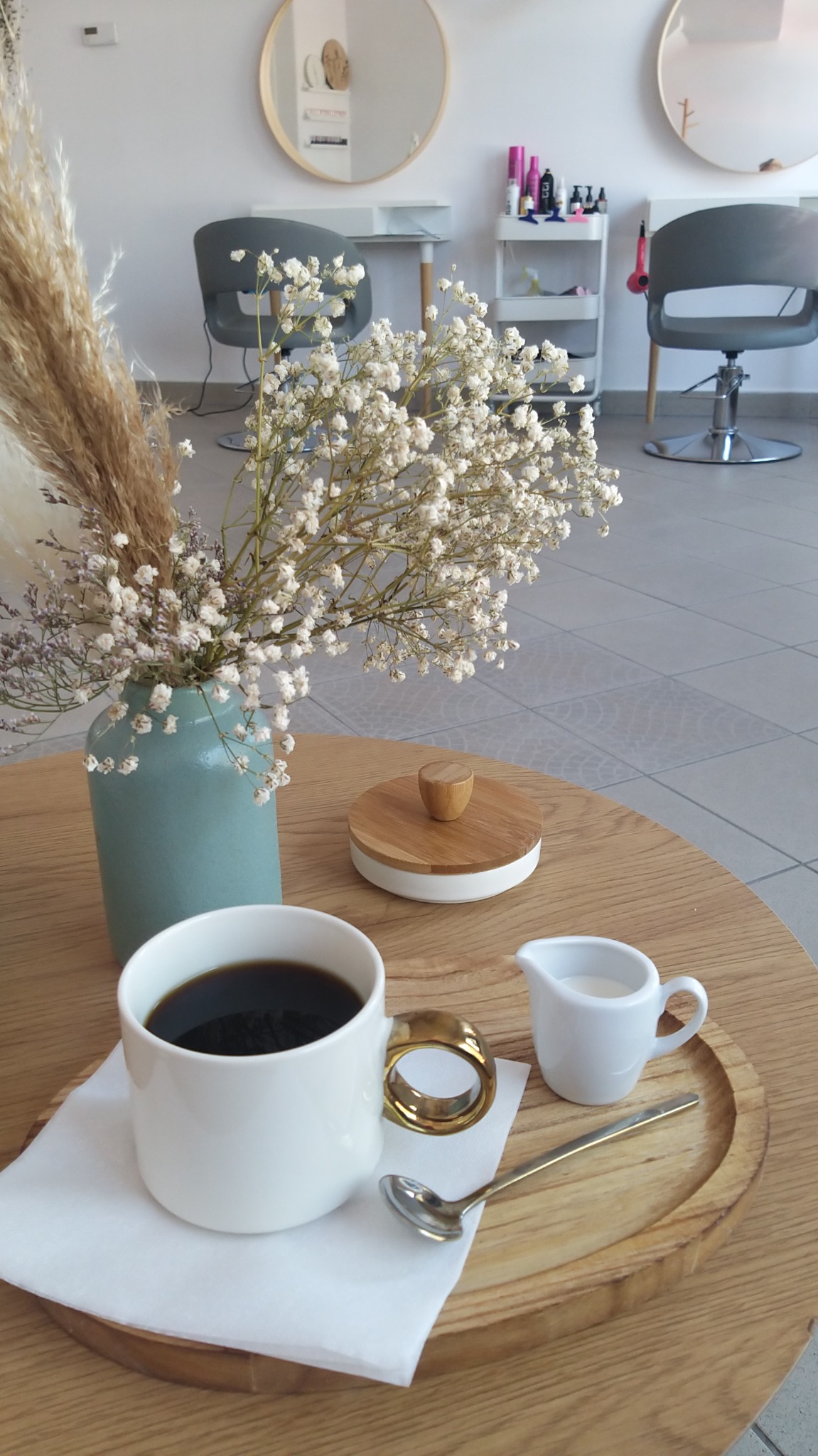 Kawa i mleko na drewnianej tacy, w tle stanowiska fryzjerskie z okrągłymi lustrami i kosmetykami, bukiet suszonych kwiatów w wazonie.