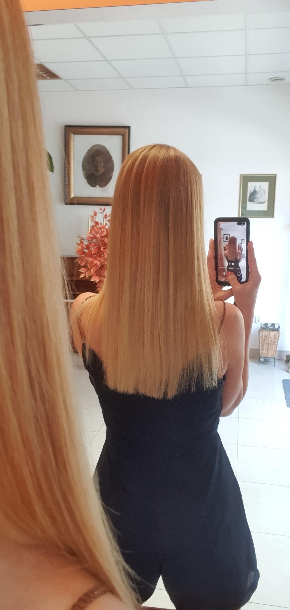 Długie, proste, blond włosy, sfotografowane od tyłu w lustrze, z widocznym odbiciem osoby robiącej zdjęcie telefonem.