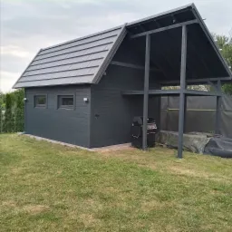 Ciemnoszara konstrukcja z zadaszeniem, łącząca zamkniętą część przypominającą mały domek z otwartą przestrzenią z grillem na trawniku.