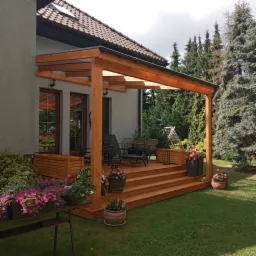 Zadaszony taras z drewna z meblami ogrodowymi i kwiatami w donicach, widok z poziomu trawnika, zieleń w tle.