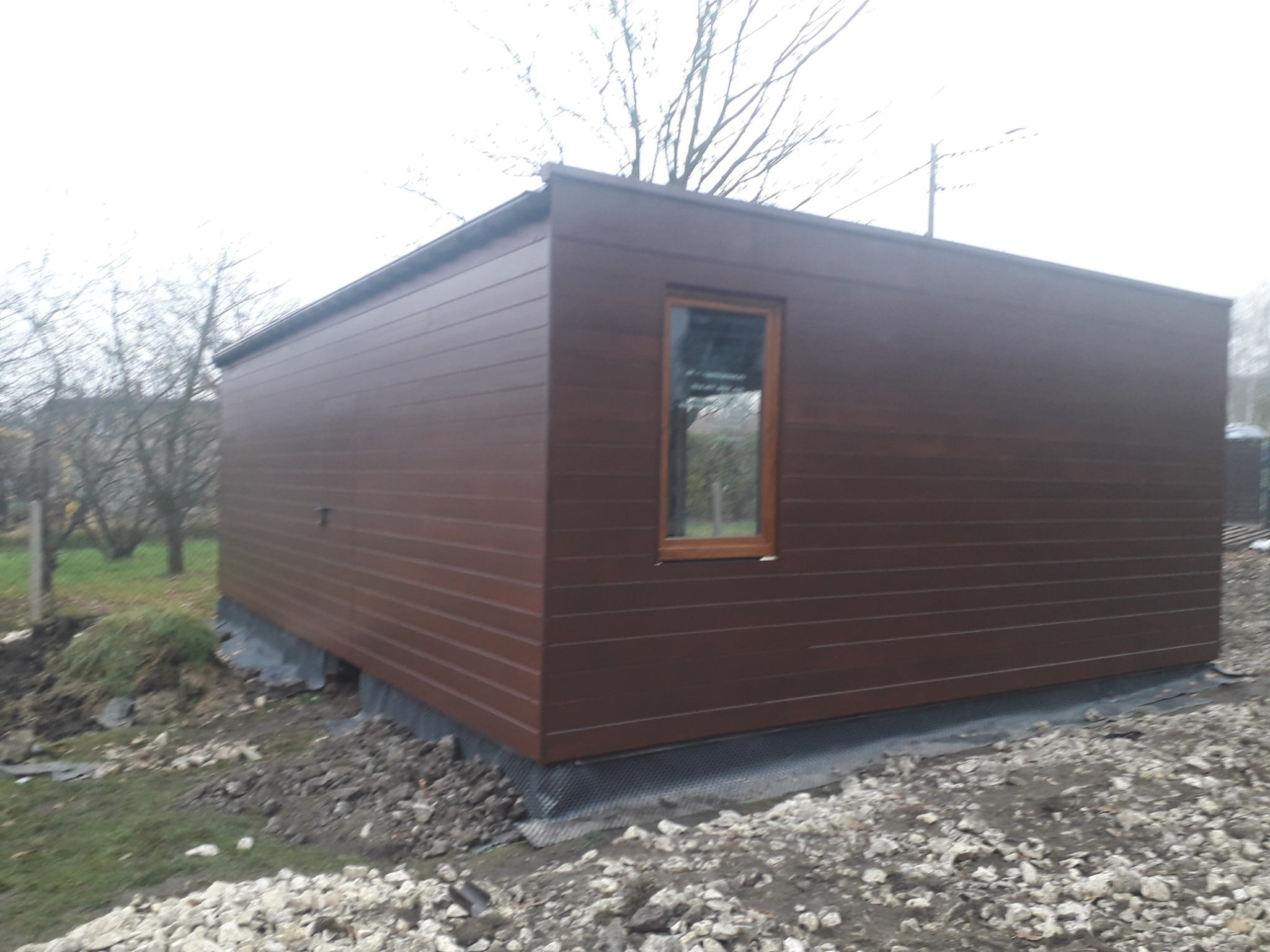 Nowoczesna, brązowa altana z poziomymi panelami, osadzona na fundamencie z kamieniami, z pojedynczym oknem z drewnianą ramą, na tle nagich drzew.