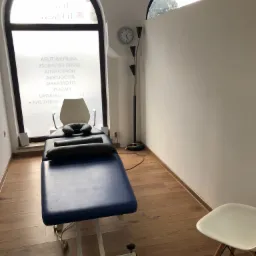 Gabinet z niebieskim łóżkiem do zabiegów, poduszkami, lampą stojącą i oknem z napisem na szkle, widok z perspektywy pacjenta.