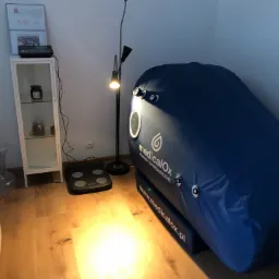 Wnętrze gabinetu z komorą hiperbaryczną MedicalOx, w tle biała szafka z przeszklonymi drzwiczkami i lampa podłogowa z regulowanymi reflektorami.
