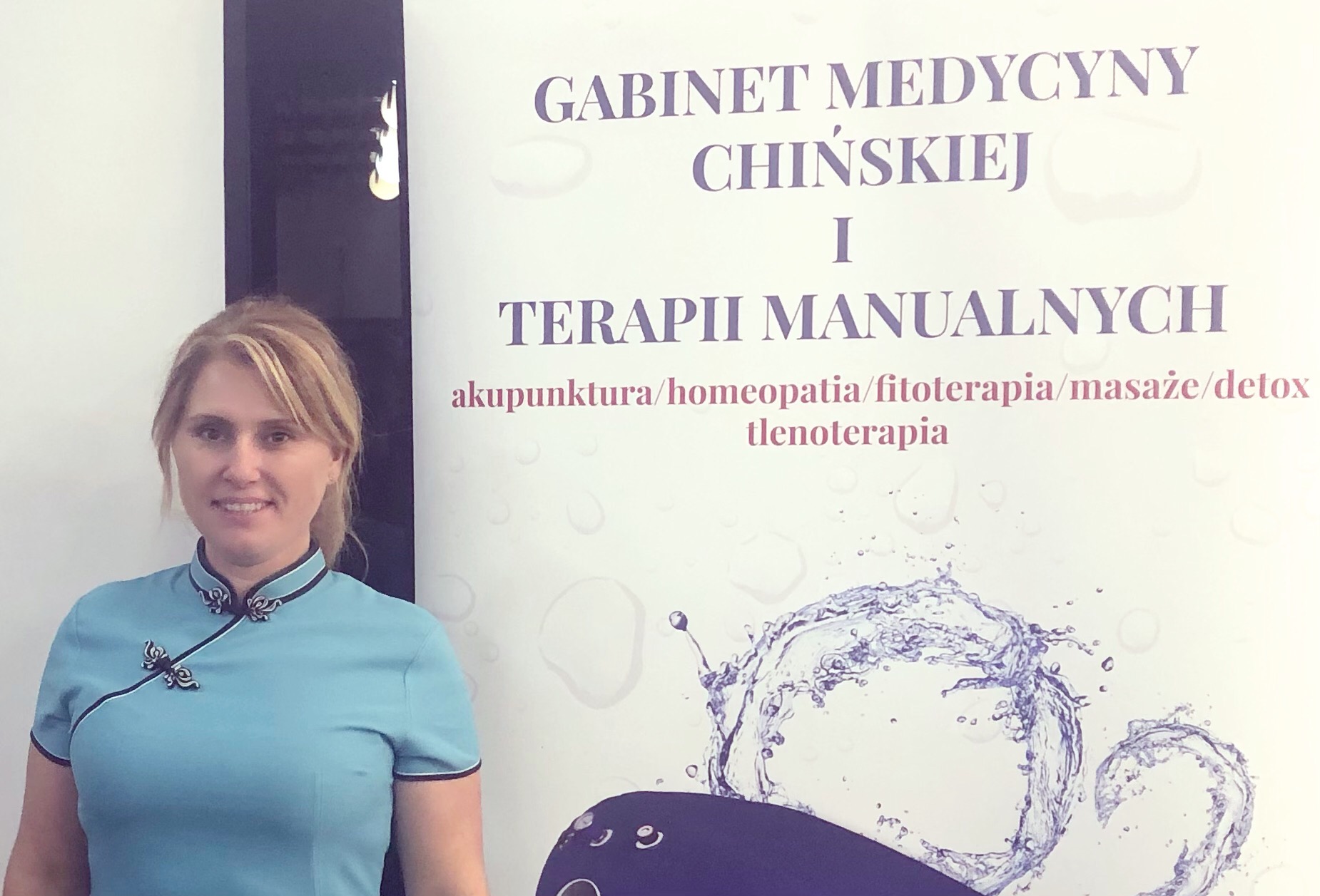 Kobieta w niebieskim uniformie z orientalnymi zdobieniami, stojąca na tle plakatu gabinetu medycyny chińskiej i terapii manualnych z wypisanymi nazwami zabiegów.