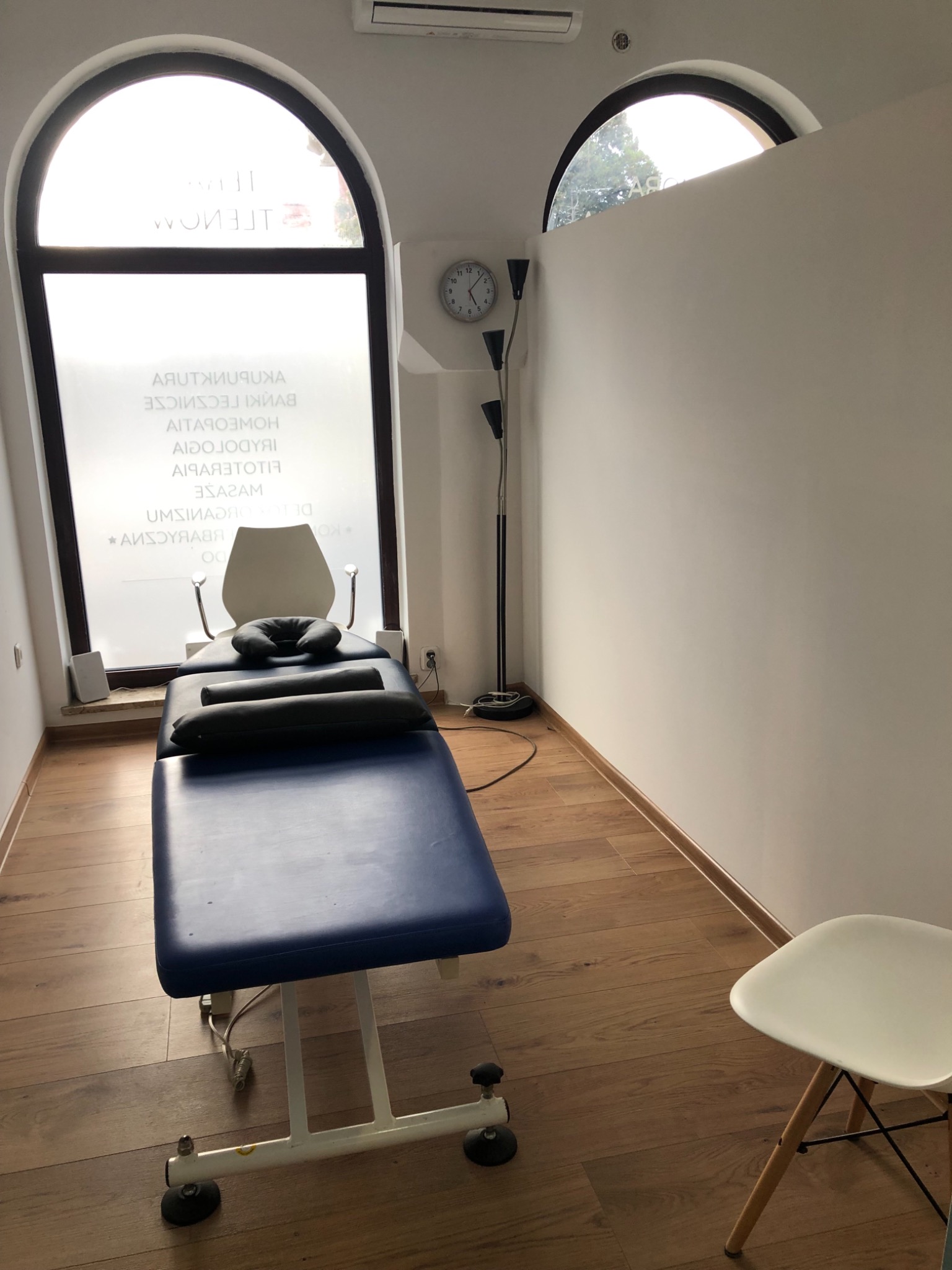 Gabinet z niebieskim łóżkiem do zabiegów, poduszkami, lampą stojącą i oknem z napisem na szkle, widok z perspektywy pacjenta.