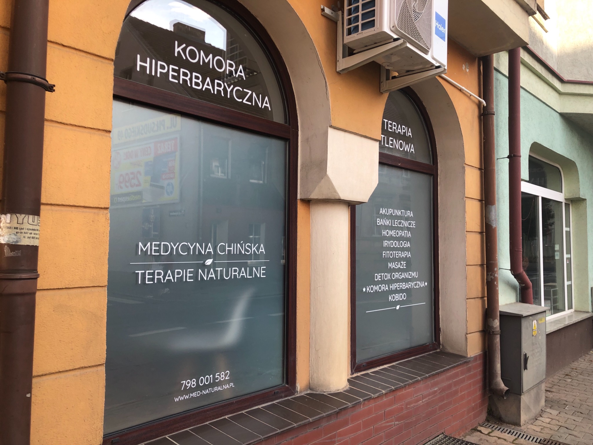 Fasada budynku z dwoma łukowatymi oknami, na których widnieją napisy: 'Komora Hiperbaryczna', 'Medycyna Chińska Terapie Naturalne' oraz 'Terapia Tlenowa' z wymienionymi zabiegami, w tym akupunktura...