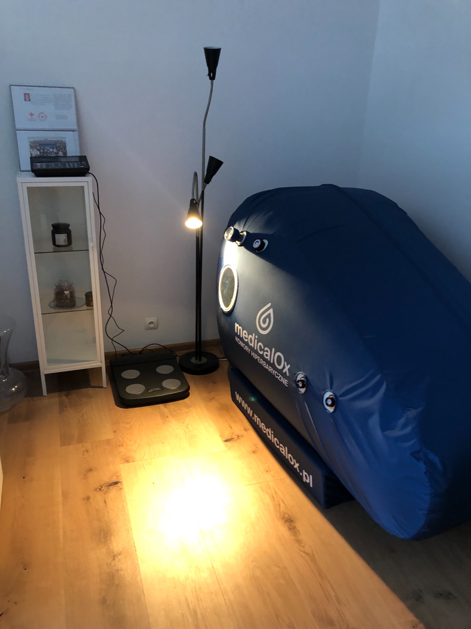 Wnętrze gabinetu z komorą hiperbaryczną MedicalOx, w tle biała szafka z przeszklonymi drzwiczkami i lampa podłogowa z regulowanymi reflektorami.