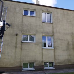 ECO-TECH Przemysław Szubert - Pracownik w kombinezonie ochronnym na drabinie spryskuje elewację budynku z użyciem opryskiwacza ciśnieniowego, widoczne zabrudzenia na ścianie.