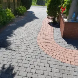 Ścieżka z kostki brukowej w odcieniach szarości i czerwieni, wijąca się obok zielonych krzewów i drzew, prowadząca do ulicy.