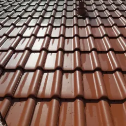 Błyszcząca, brązowa dachówka ceramiczna widziana pod kątem, z widocznym kominem wentylacyjnym i kroplami wody po deszczu lub myciu.