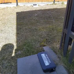 Czarny karmnik deratyzacyjny z napisem 'NIE DOTYKAĆ' umieszczony na szarej betonowej płycie na tle trawnika i drewnianego ogrodzenia.