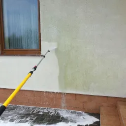 Czyszczenie elewacji domu z widocznym efektem przed i po, przy użyciu lancy teleskopowej z dyszą rozpylającą wodę pod ciśnieniem, w tle okno.