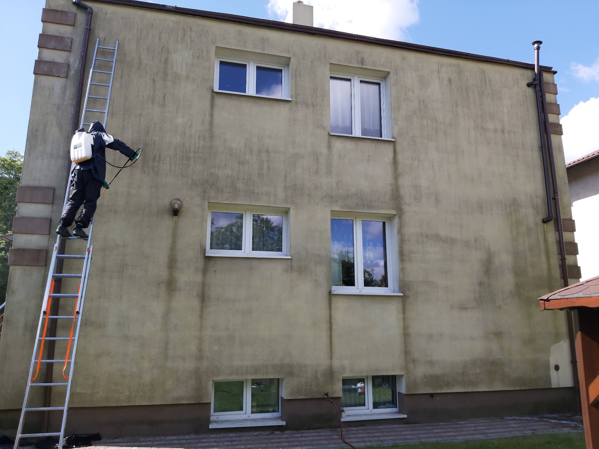 Pracownik w kombinezonie ochronnym na drabinie spryskuje elewację budynku z użyciem opryskiwacza ciśnieniowego, widoczne zabrudzenia na ścianie.